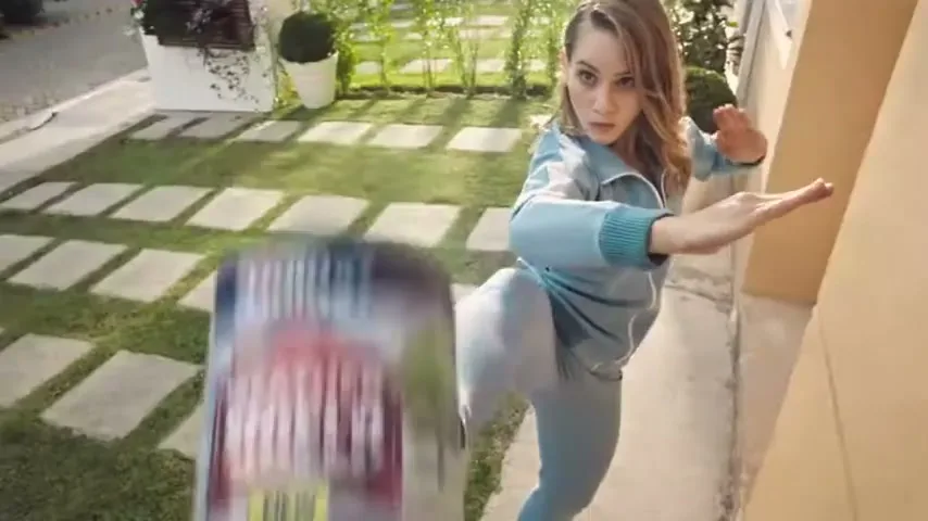 A young woman in a blue tracksuit kicks a large can of Dulux Weathershield paint in a sunny garden.
