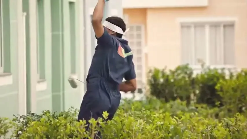A male painter in a blue uniform and white headband paints the exterior wall of a light green building with a roller, surrounded by green bushes.
