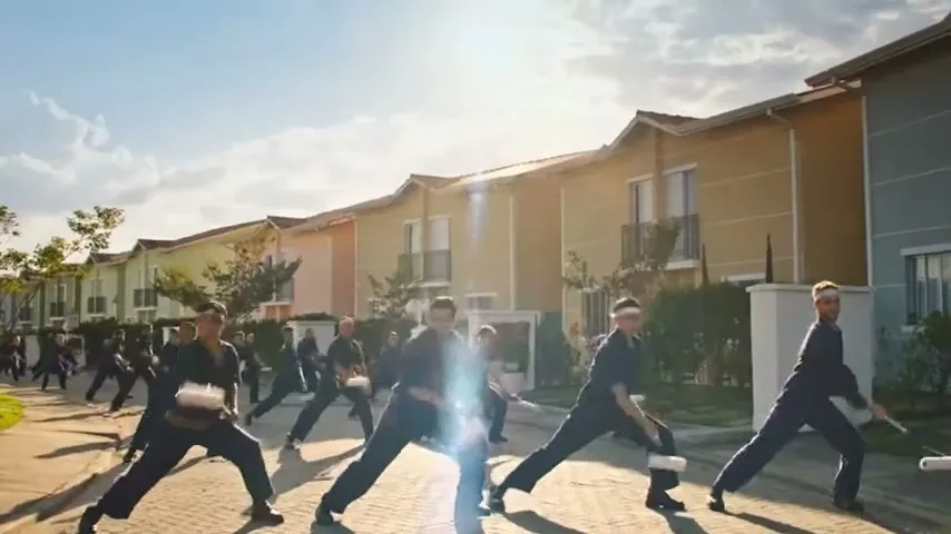 A large group of men in dark uniforms and headbands perform synchronized martial arts moves with paint rollers on a sunny suburban street.