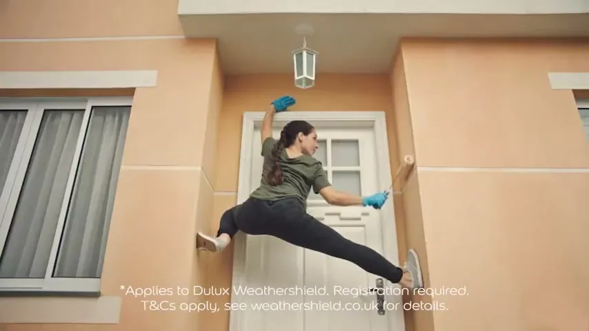 A woman in a green shirt and blue gloves is painting the exterior wall of a house while doing a split between two walls.