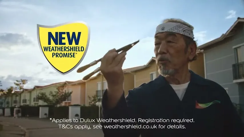 An older man in a bandana and work shirt holds up paint brushes like chopsticks, smiling in front of colorful houses under a bright sky.