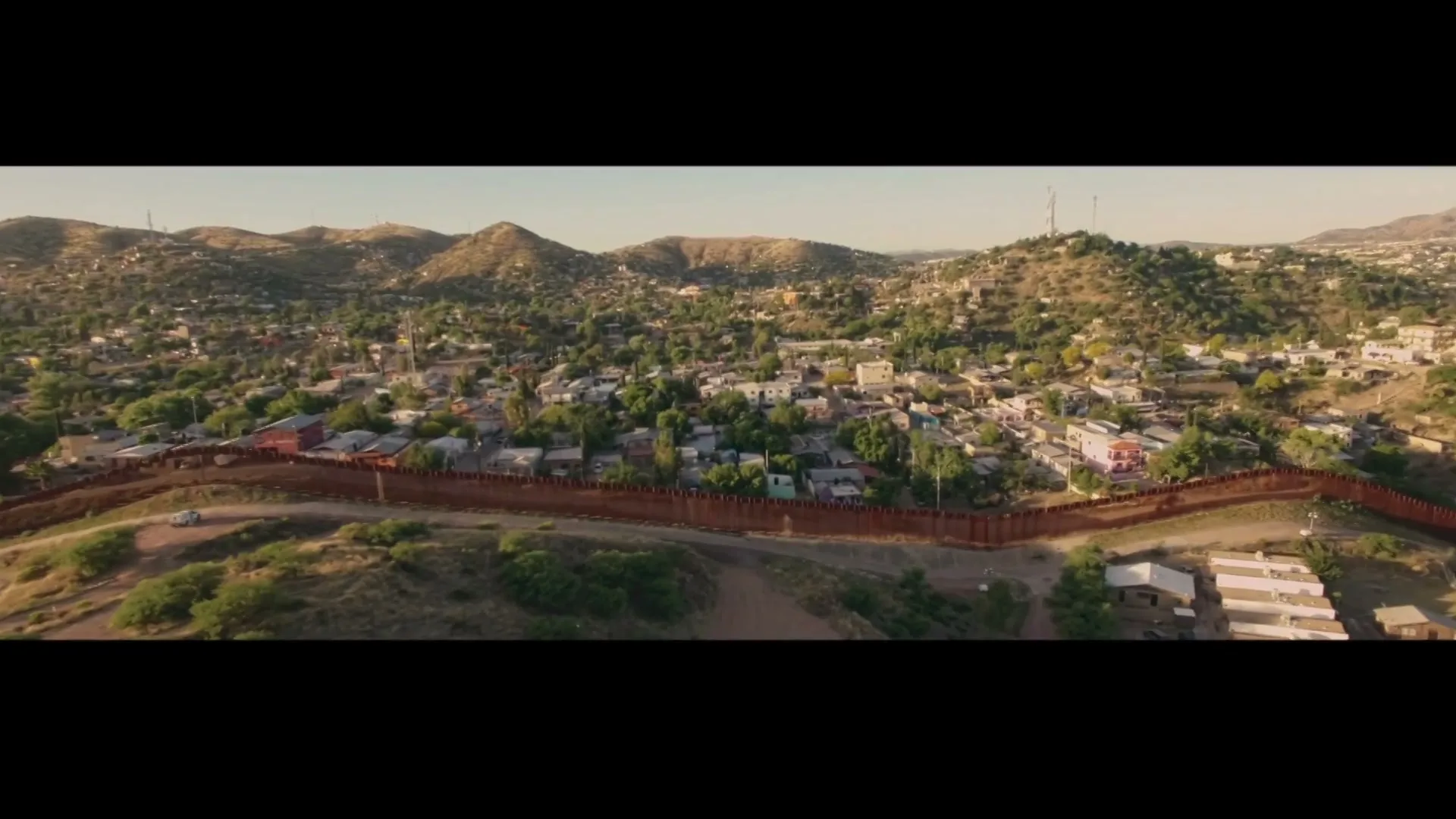 An aerial wide shot shows a long, rusty border wall dividing a town nestled in a valley between hills under a clear sky.