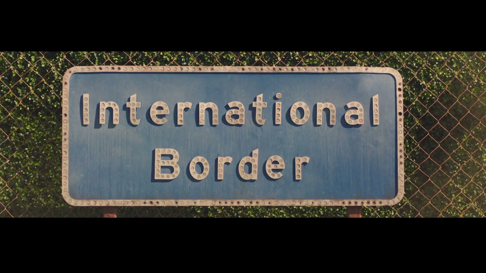A blue "International Border" sign with studded letters is seen against a chain-link fence and green foliage.