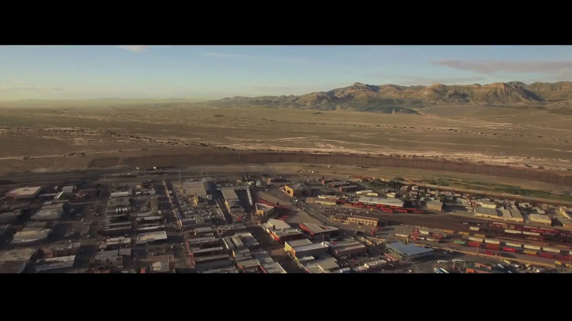 An aerial view shows a small town with a long, brown wall separating it from a vast desert landscape under a clear sky.