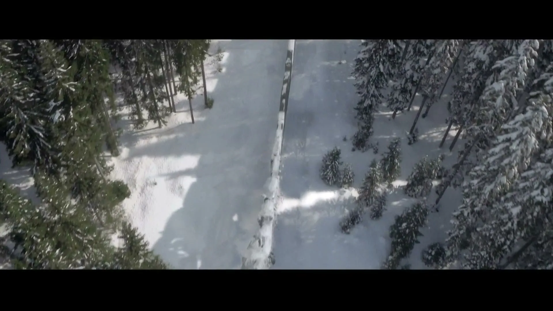 An aerial view shows a bobsled track winding through a snow-covered forest on a bright, sunny day.