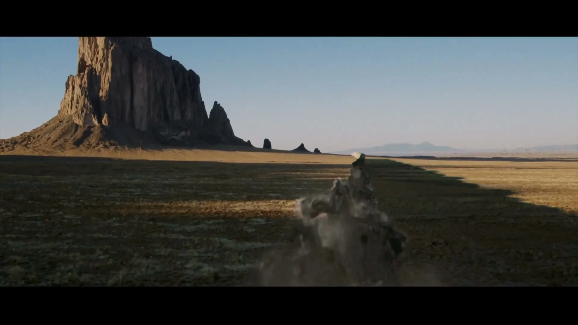 A long wall of rocks and dirt crumbles and falls apart in a vast desert landscape with a large mesa in the background under a clear blue sky.
