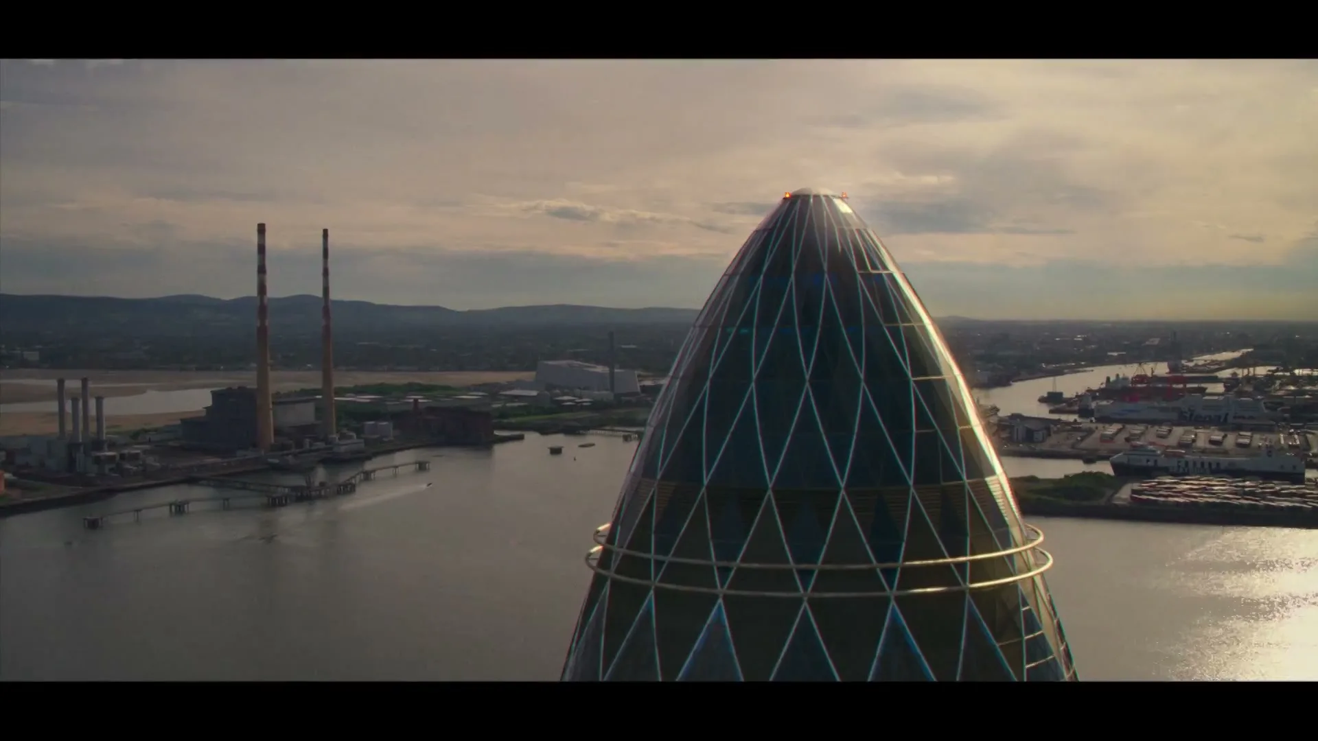 An aerial shot of a modern, glass-paneled building with a conical top, overlooking a river with industrial structures and a city skyline under a cloudy sky.