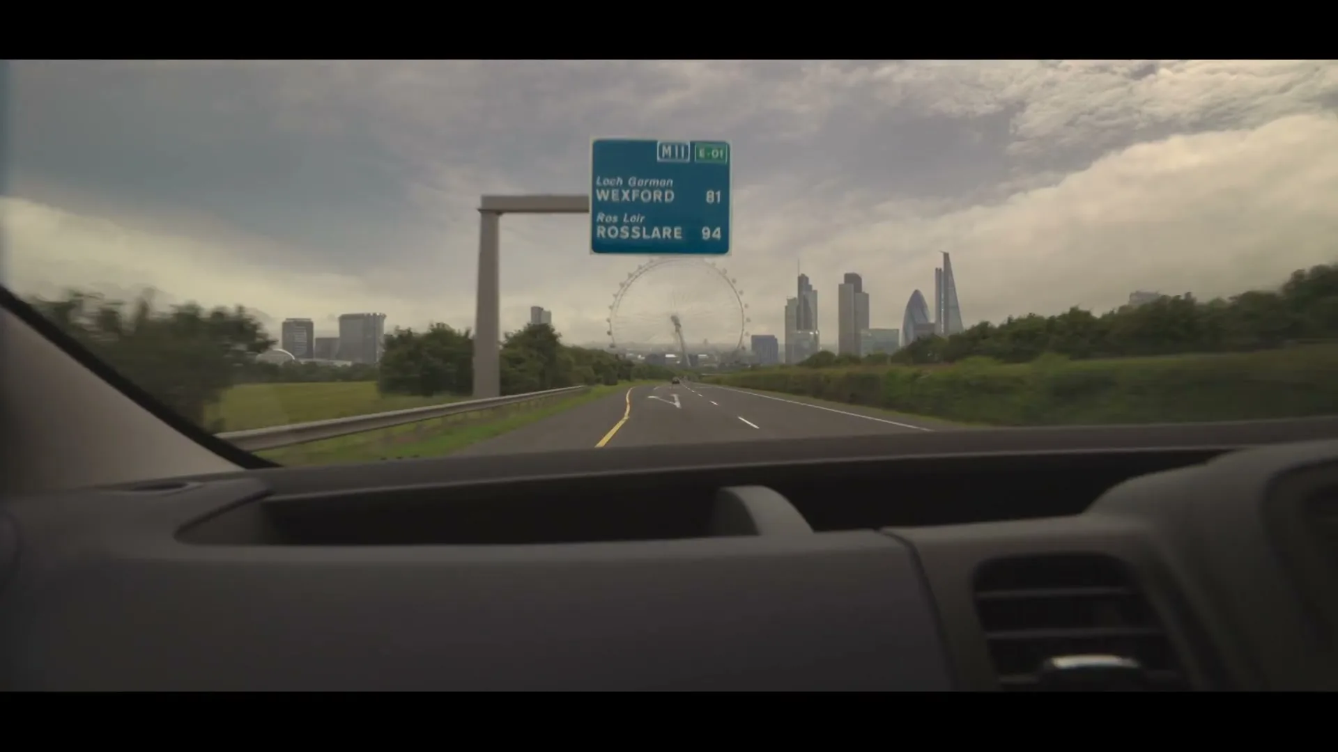A POV shot from inside a moving car shows a highway leading towards a cityscape with a Ferris wheel and skyscrapers under a cloudy sky.