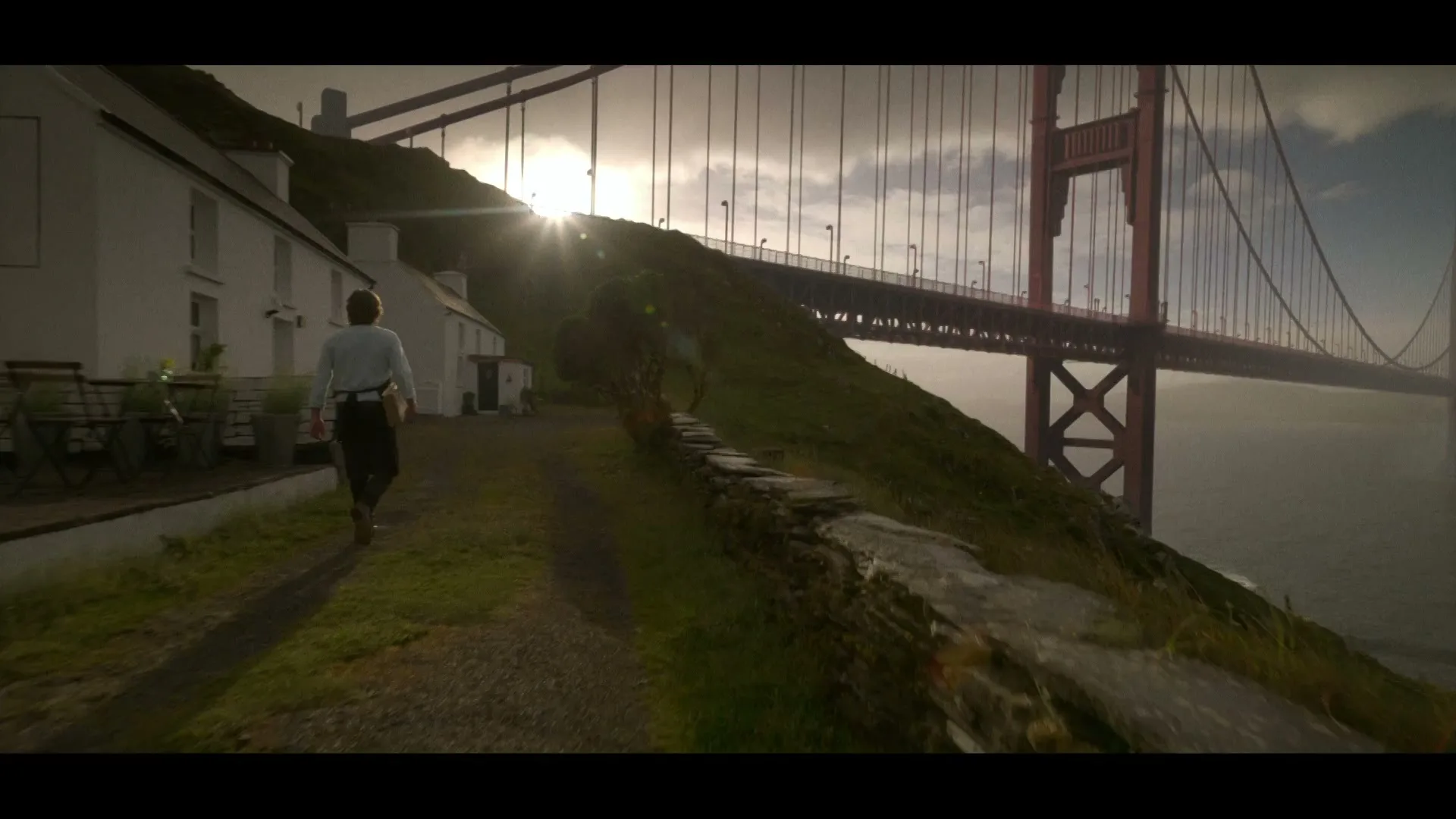 A man walks away from a white house on a grassy hill towards the Golden Gate Bridge at sunset, with a stone wall on the right.