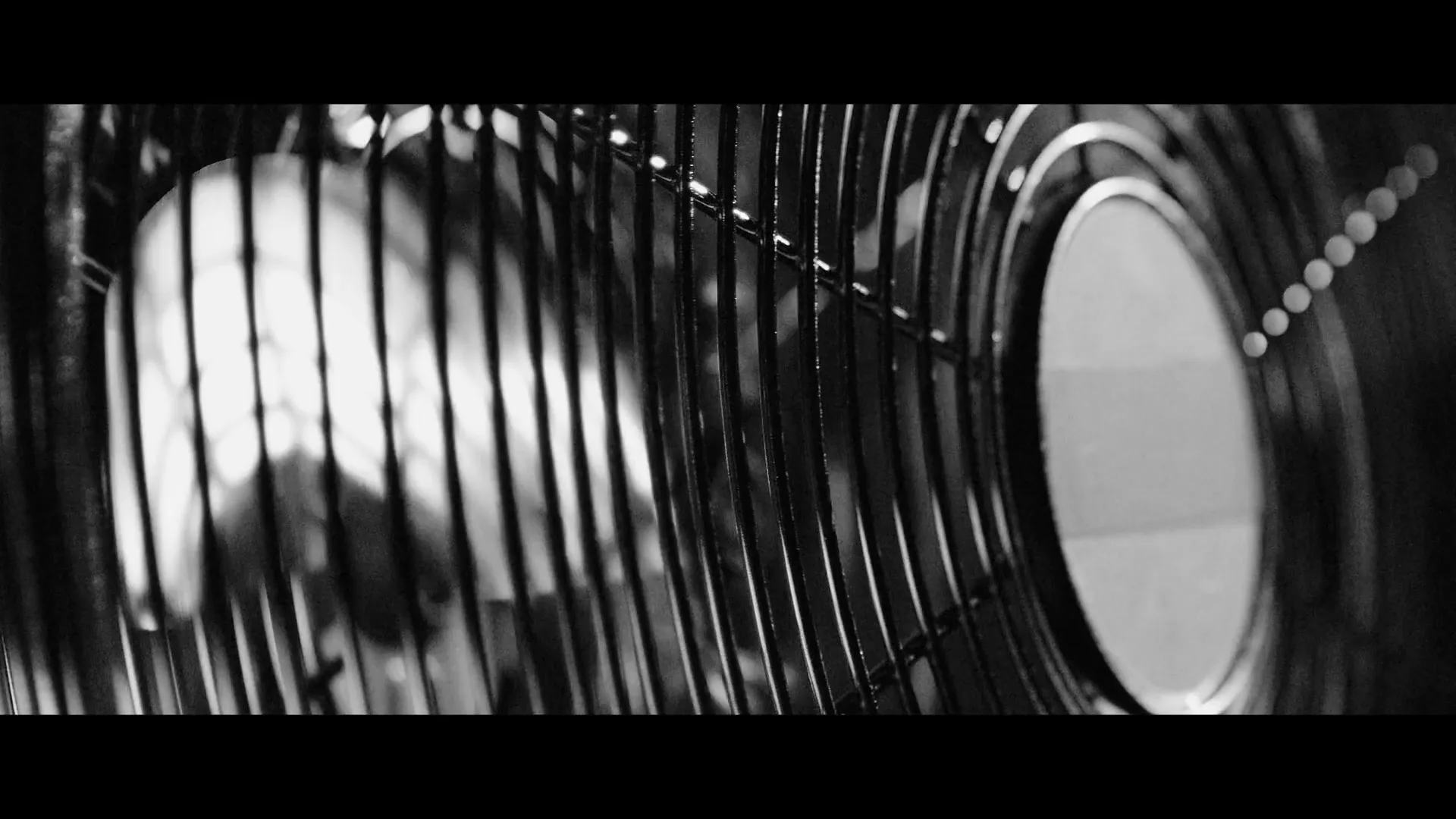 A black and white extreme closeup shot of a fan's grill, with a blurry figure visible behind it, in an interior setting.