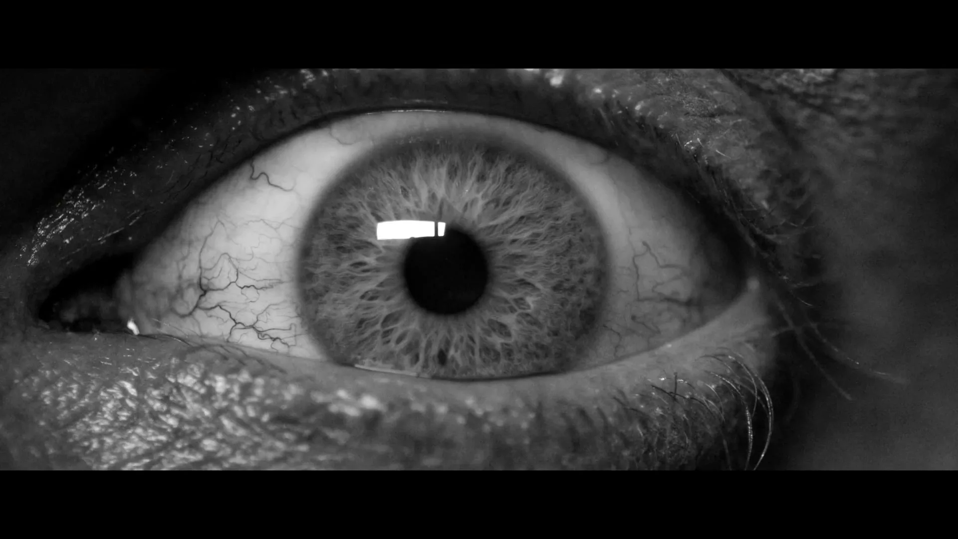 A black and white extreme closeup of a person's eye opening, revealing a bloodshot iris, in a dark setting.