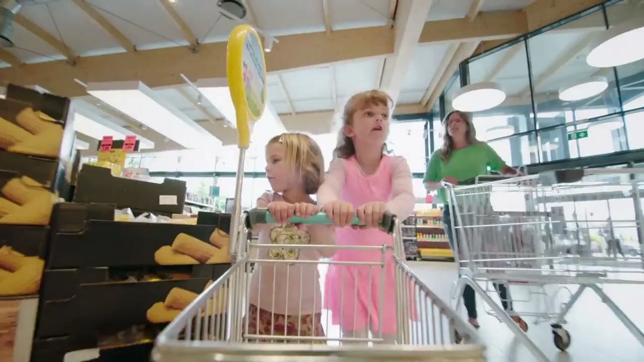 Two young girls push a shopping trolley through a supermarket aisle, with an adult woman pushing another trolley behind them.