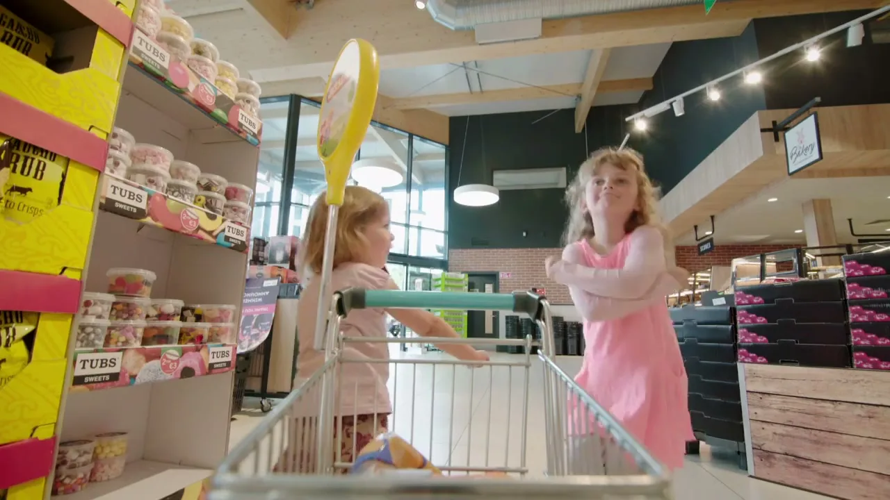 Two young girls, one in a pink dress and the other in a floral skirt, are in a supermarket aisle, looking at sweets from a low-angle shot from inside a shopping trolley.