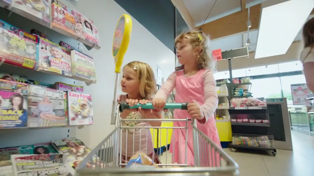 Two young girls, one in a pink dress and one in a pink top, push a shopping trolley down an aisle in a supermarket.