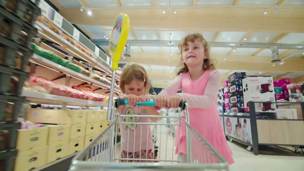 Two young girls push a shopping trolley through a supermarket aisle, looking around with curiosity and excitement.