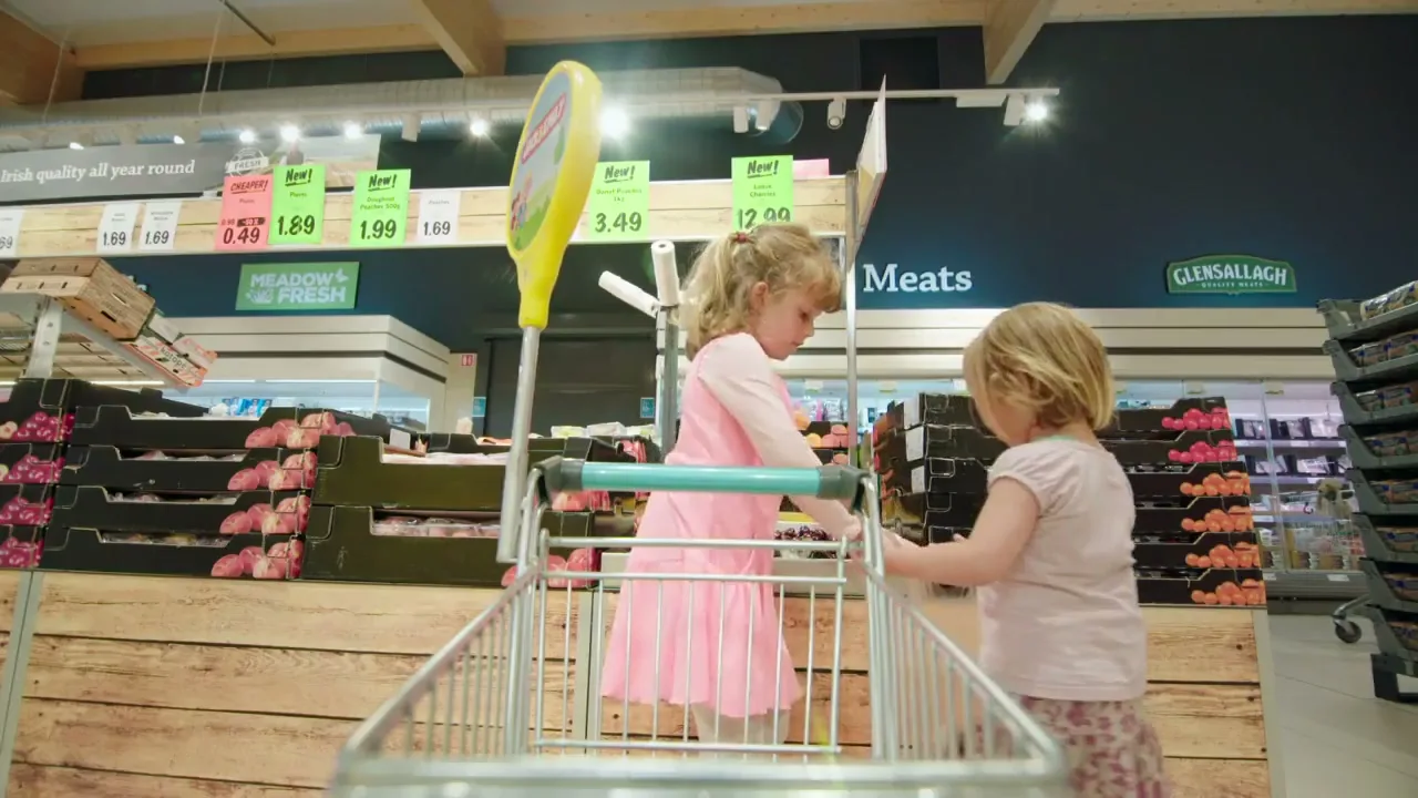 Two young girls in a supermarket aisle, one in a pink dress and the other in a white t-shirt, are putting fruit into a shopping trolley.