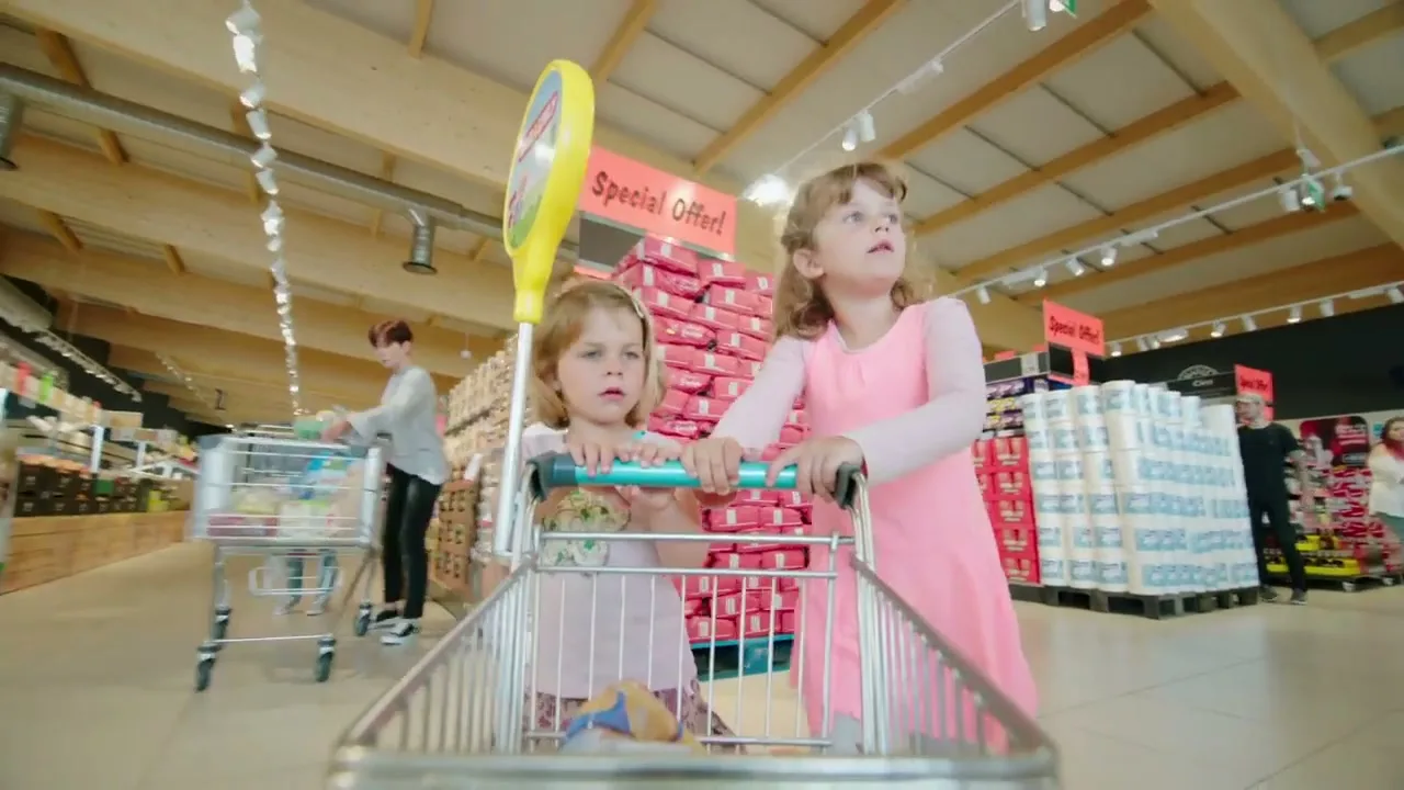 Two young girls, one in a pink dress and the other in a floral top, are riding in a shopping cart through a brightly lit supermarket aisle, looking around with curiosity.