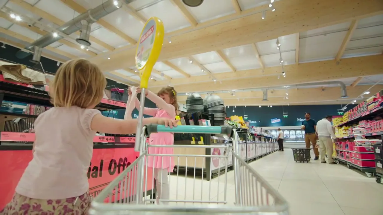 Two young girls push a shopping trolley through a brightly lit supermarket aisle, with other shoppers visible in the background.