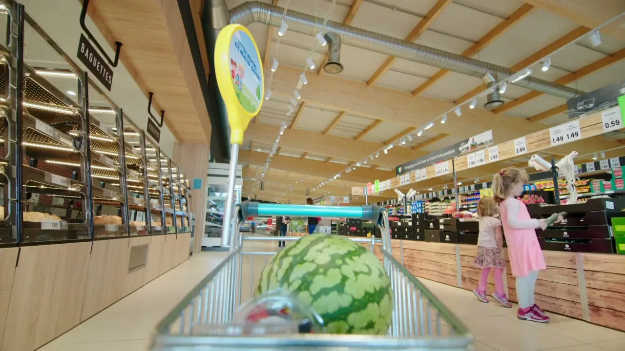 Two young girls in a supermarket aisle, one holding a cucumber, as seen from inside a shopping trolley with a watermelon.