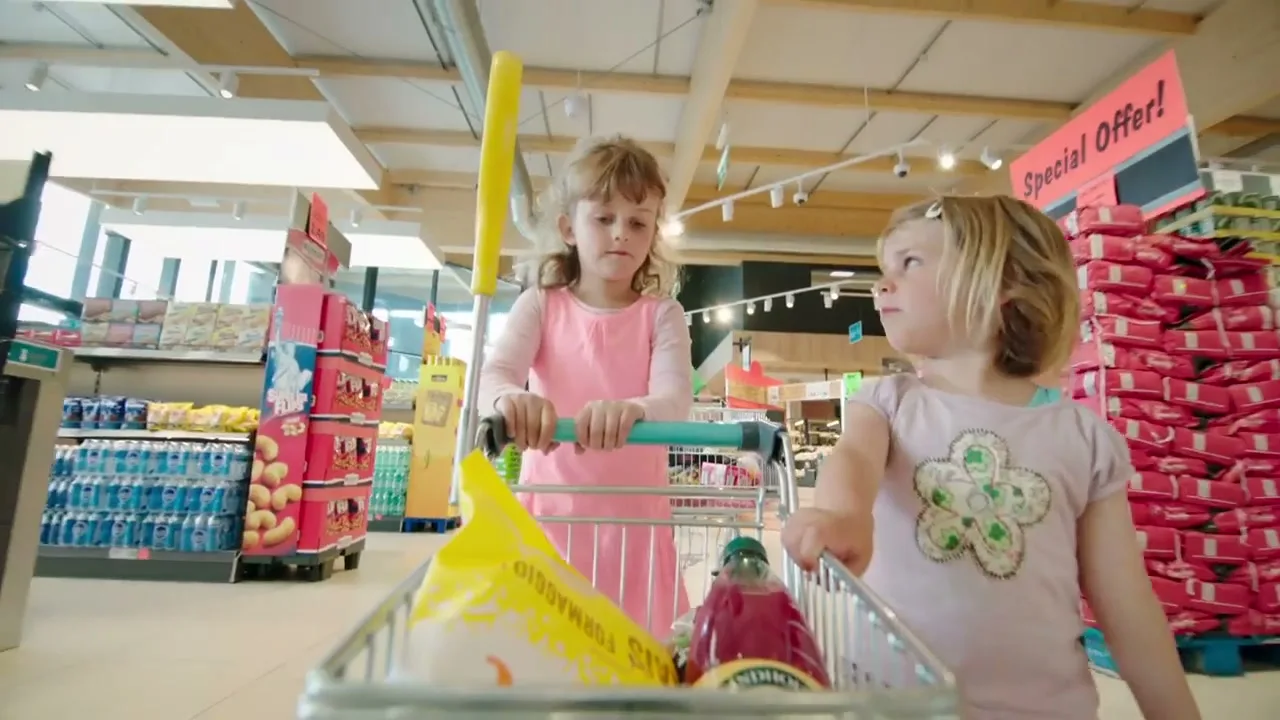 Two young girls, one in pink and one in a light t-shirt, are pushing a shopping cart through a brightly lit supermarket aisle.