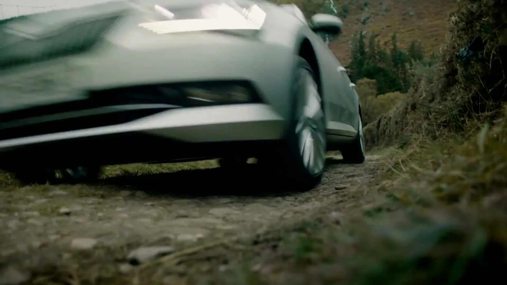 A silver Skoda car drives towards the camera on a narrow, rocky dirt road in a rural, mountainous landscape.