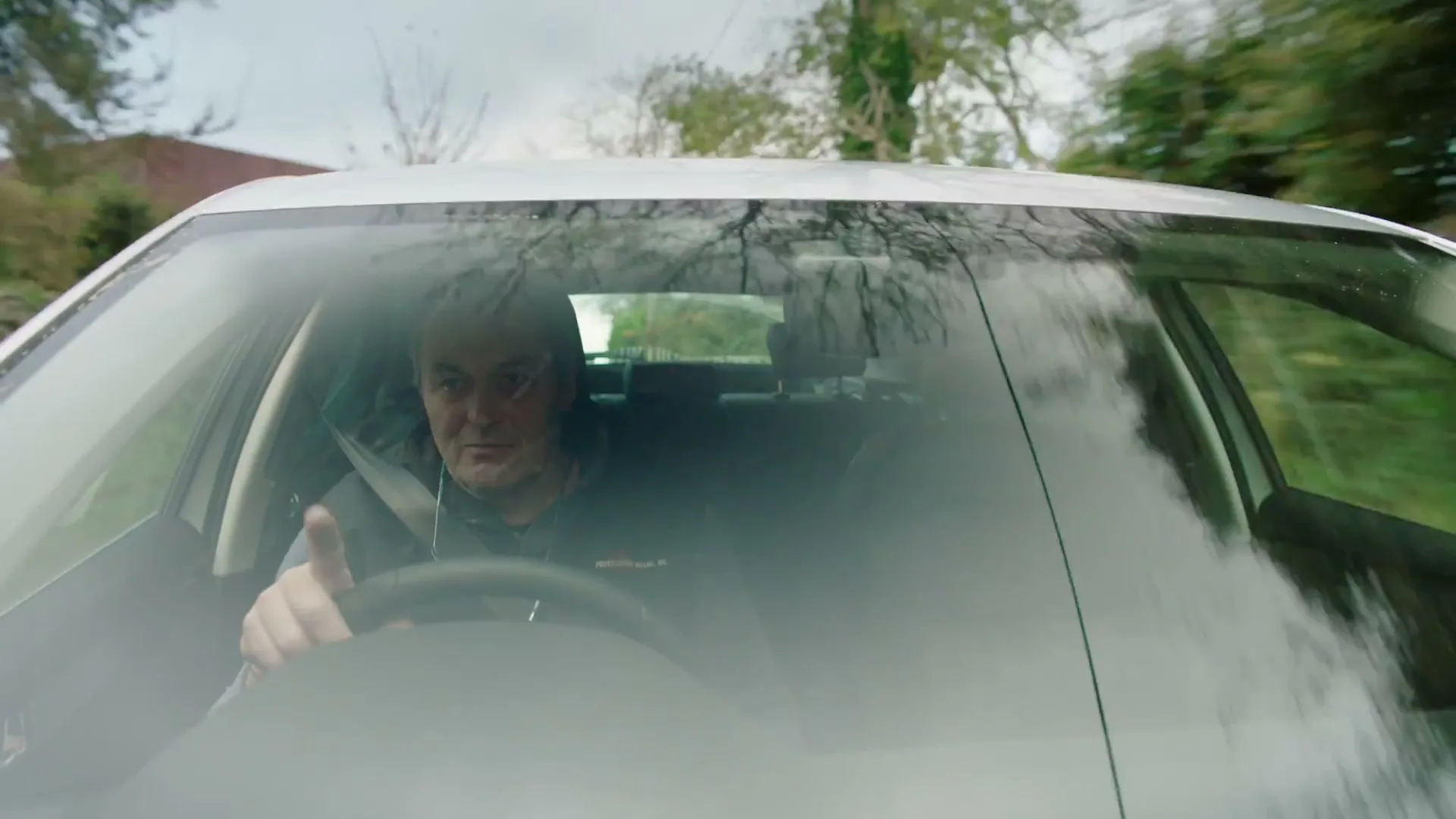 A man in his 50s drives a white Skoda car on a country road, seen through the windshield.