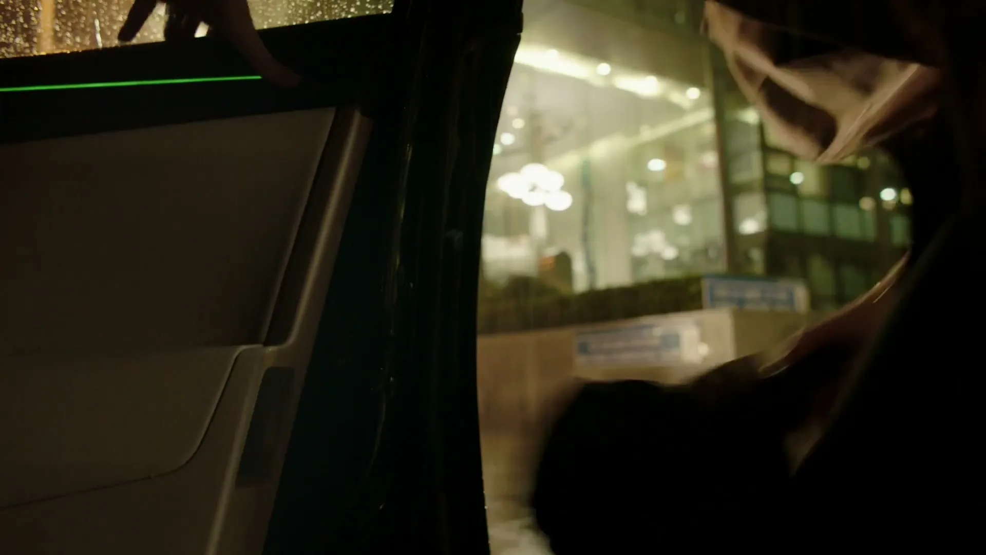 A person in a trench coat closes a car door in the rain at night, with a modern building in the background.