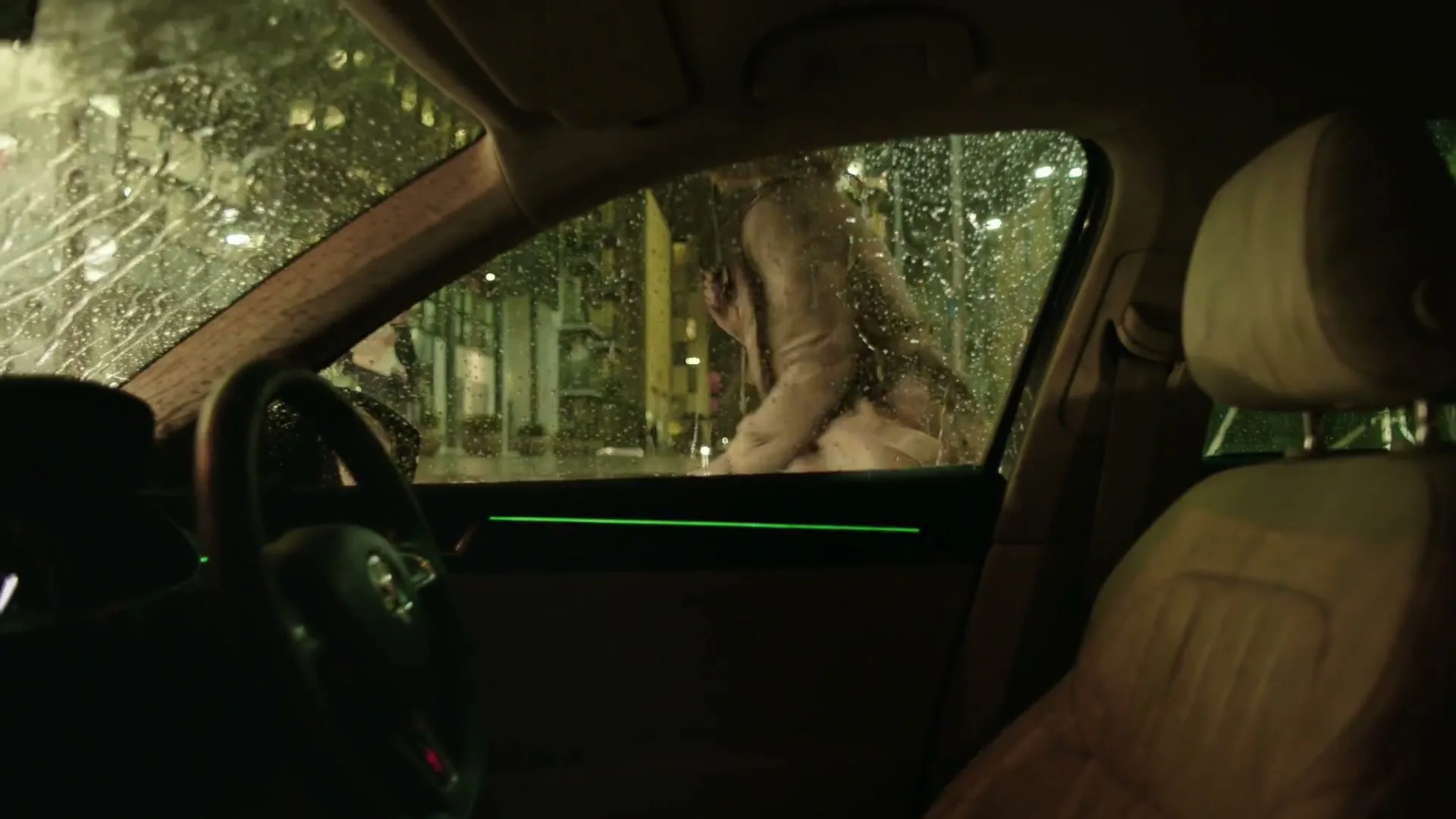 A person in a trench coat walks past a car window, which is covered in raindrops, on a dark, rainy street at night.