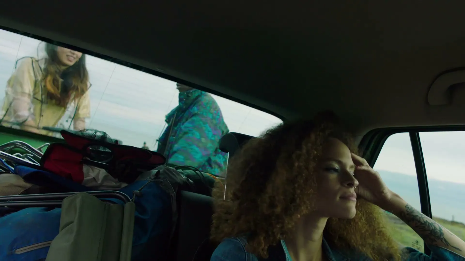 A young woman with curly hair sits in the passenger seat of a car, looking out the window as two other people load luggage into the back.