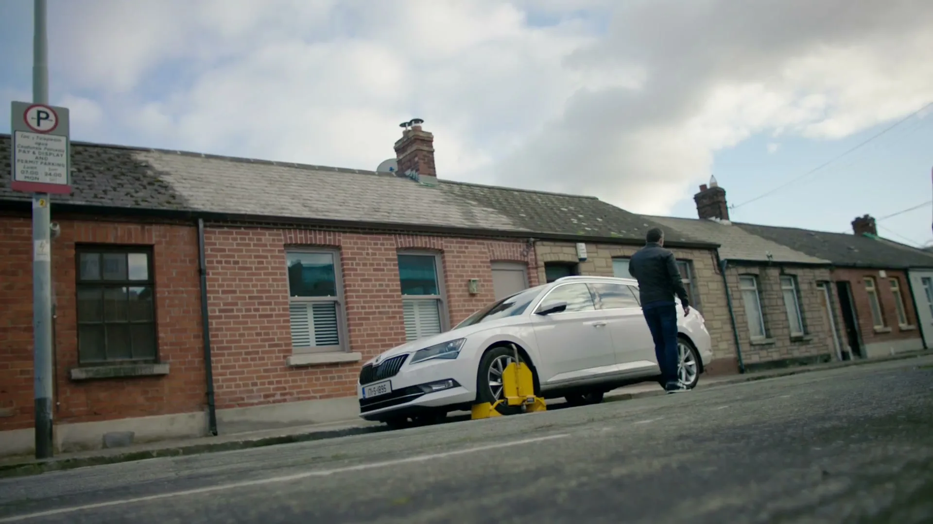 A man in a dark jacket and jeans walks towards a white Skoda car with a yellow wheel clamp on a residential street in Ireland during the day.