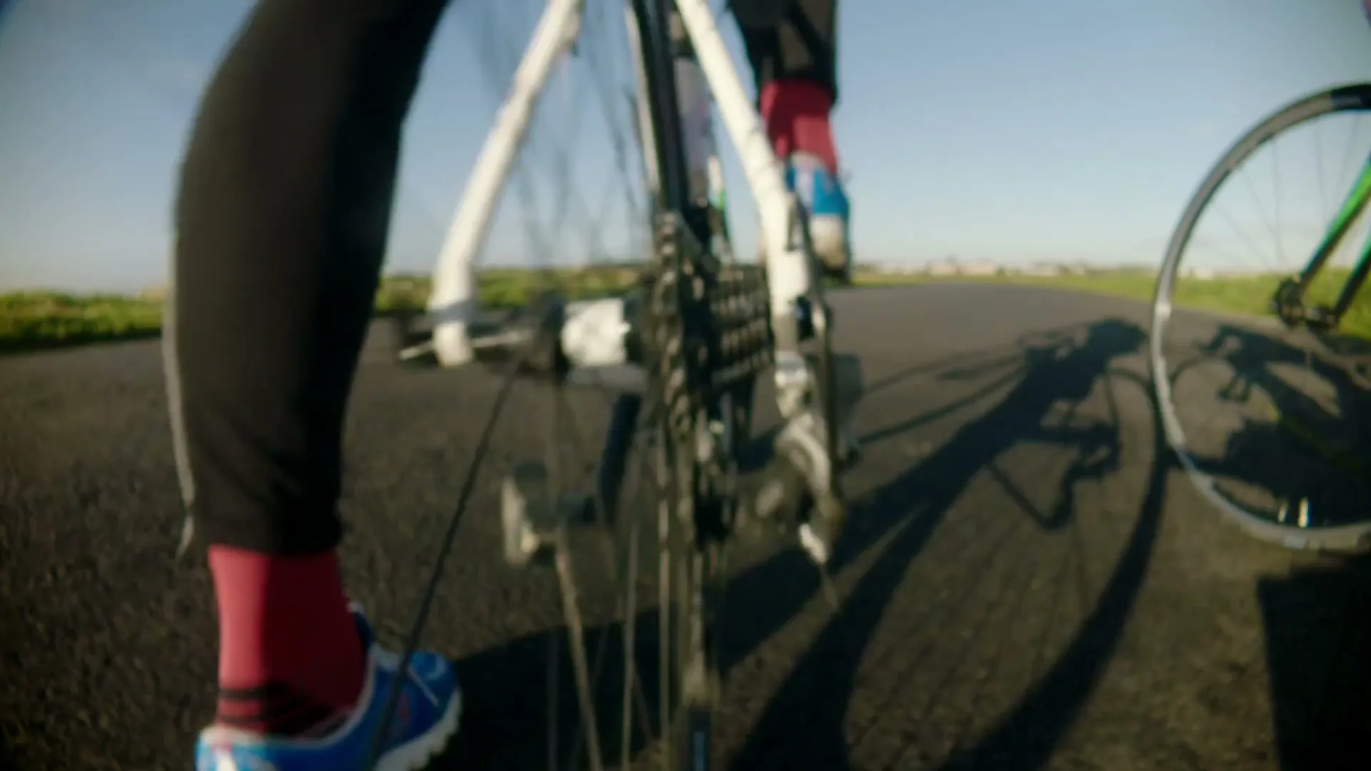 Two cyclists ride their bikes on a paved road, with a close-up on the rear wheel and gears of one bike.