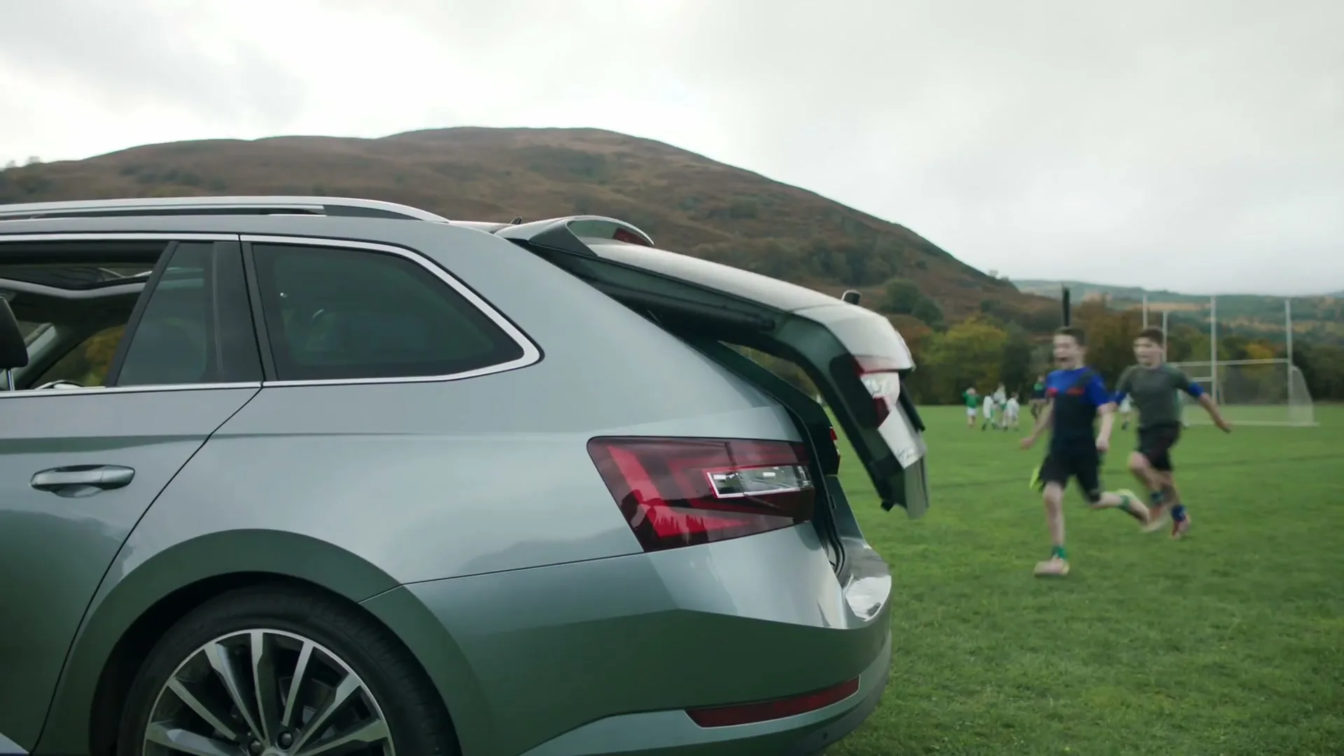 A grey Skoda Superb Combi with its boot open is parked on a grassy field as two boys run past, one carrying a rugby ball.