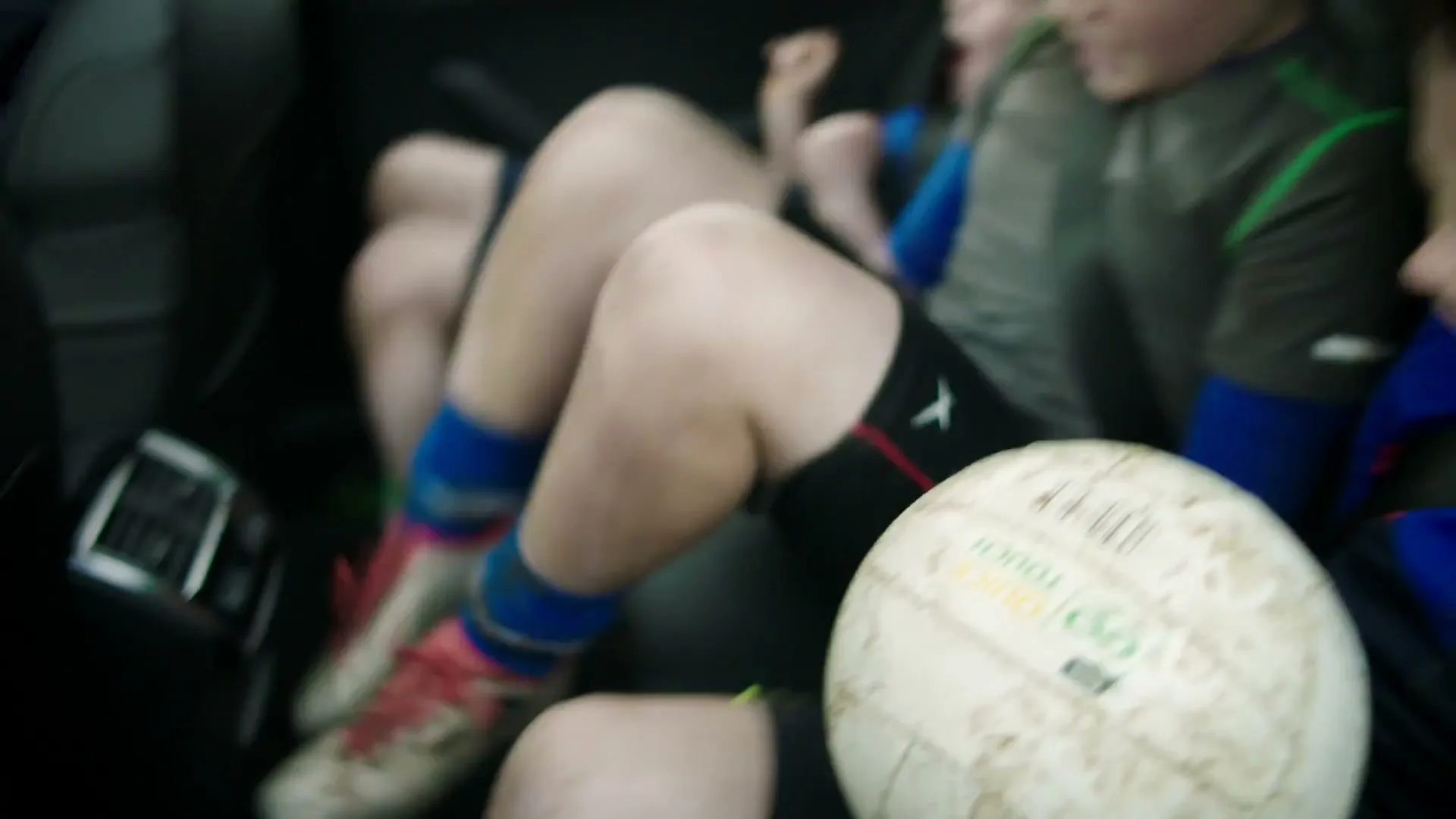 Three young boys in sports gear are crammed in the back seat of a car, with a football visible in the foreground.