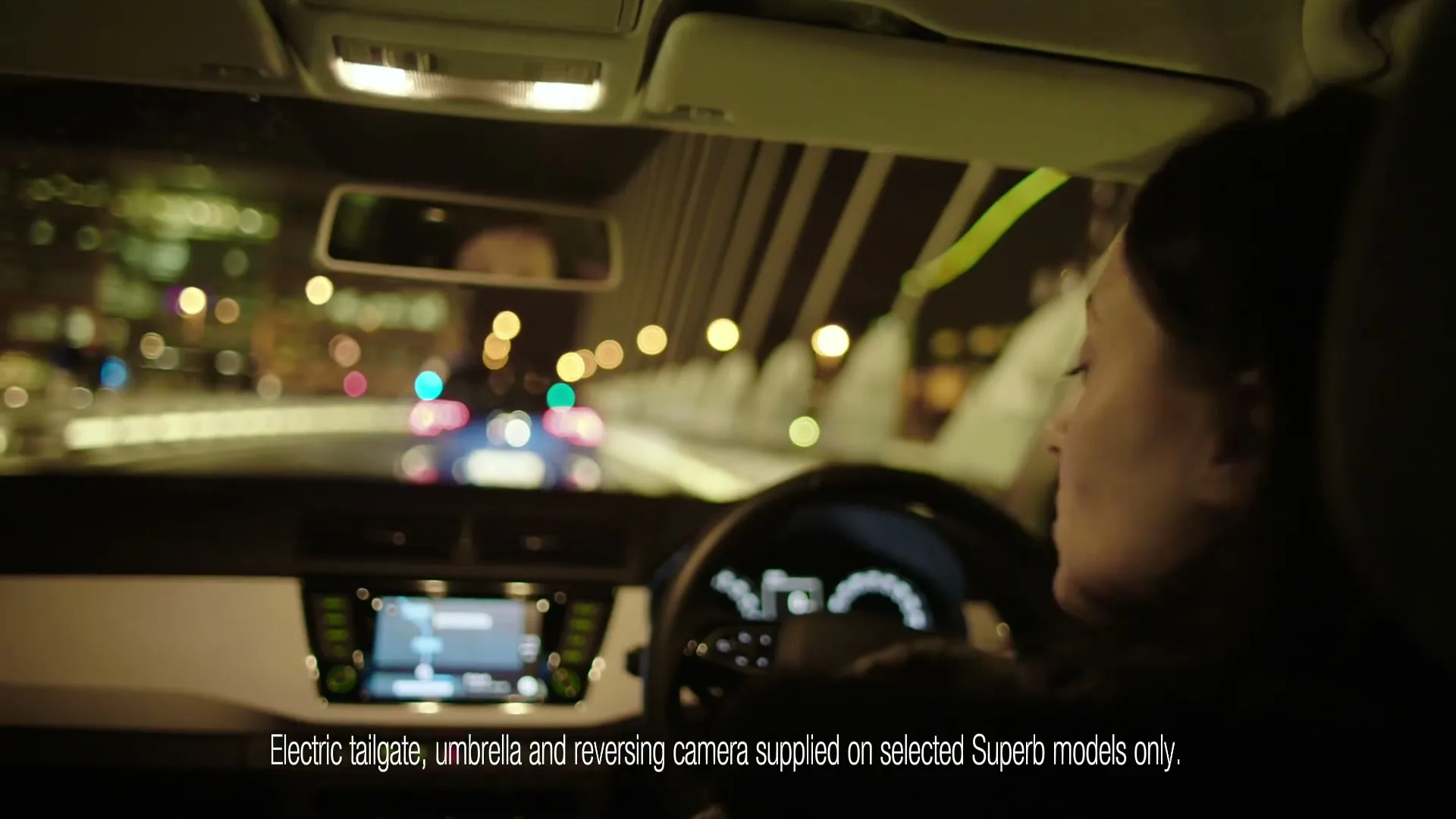 A woman with dark hair drives a car at night, with city lights blurred in the background, and text on screen.
