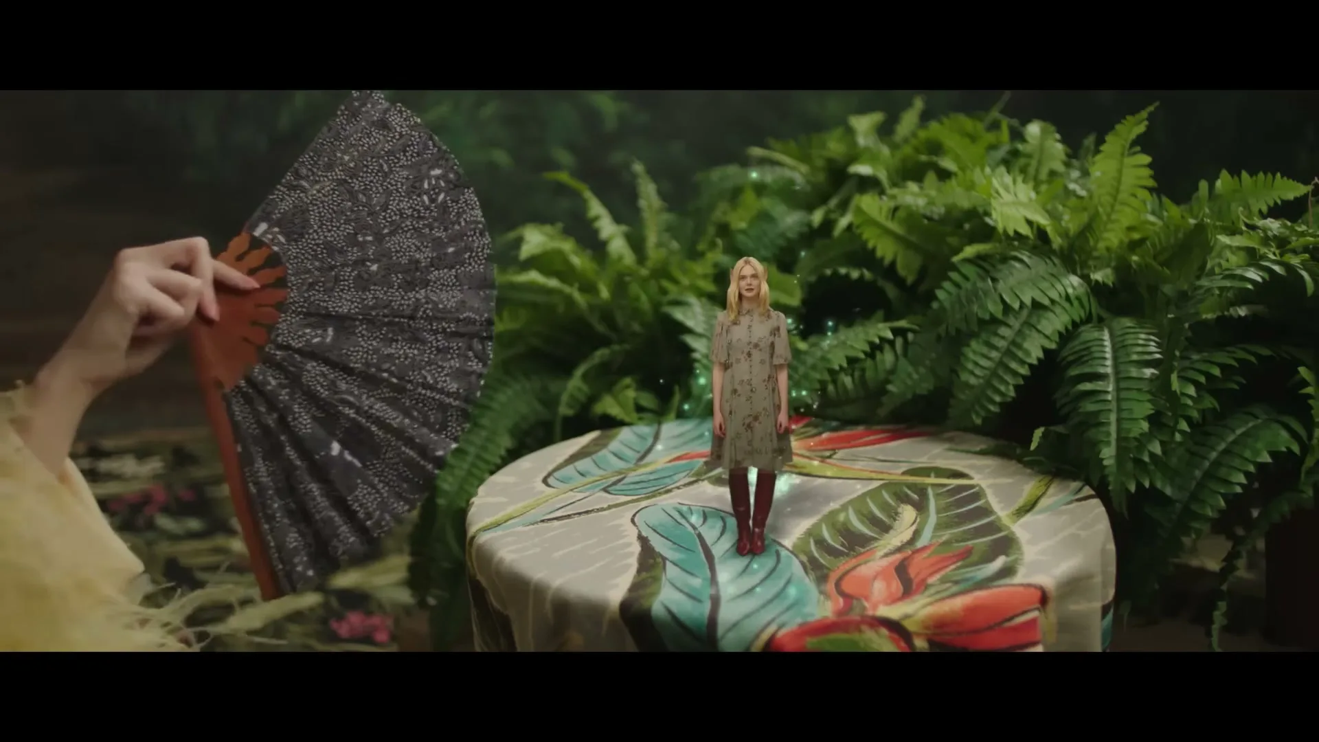 A tiny Elle Fanning stands on a patterned table, surrounded by lush green plants, as a hand holding a fan appears on the left.