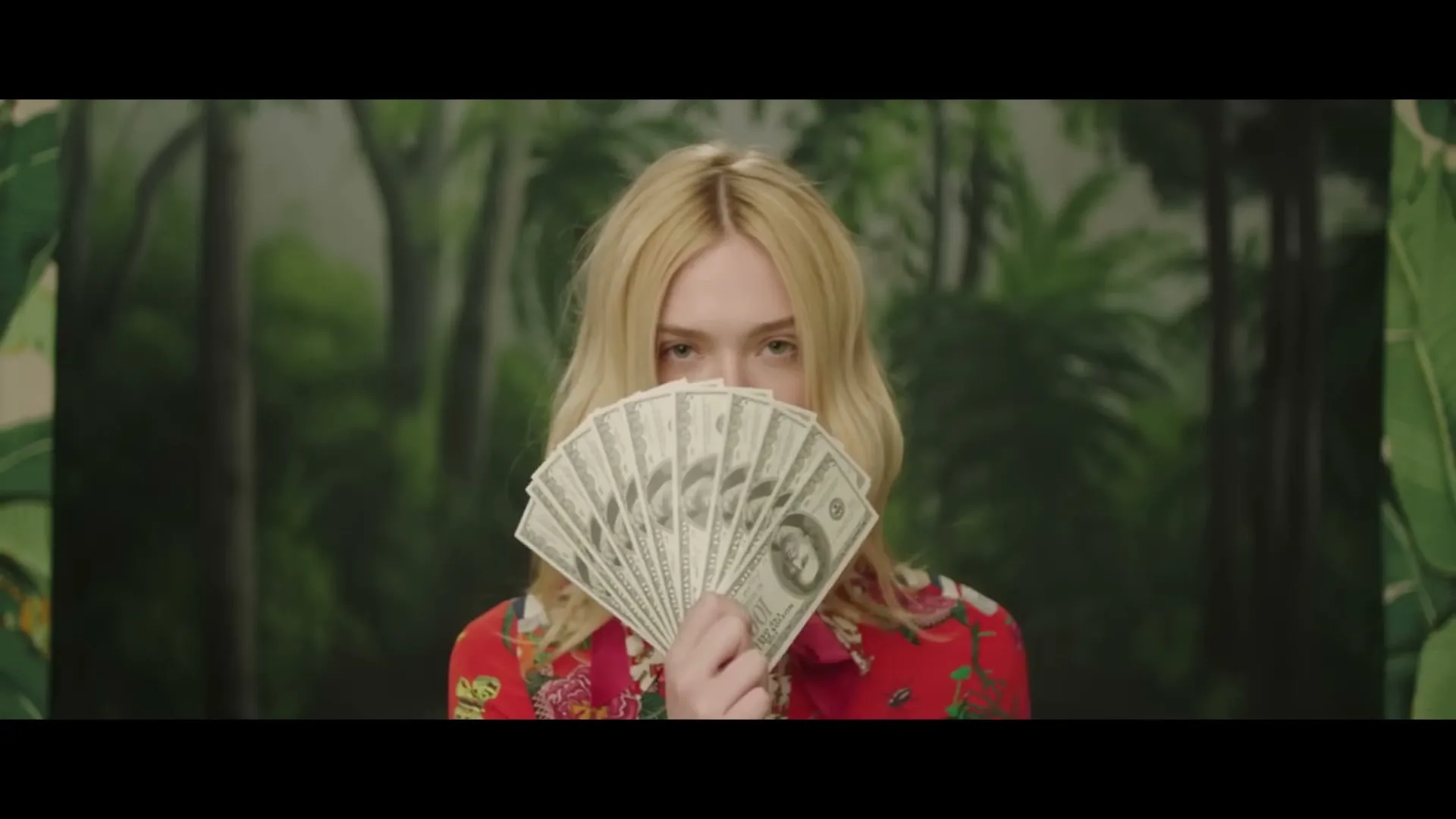 Elle Fanning, a young blonde woman, holds a fan of money in front of her face, looking directly at the camera in a studio with a jungle backdrop.