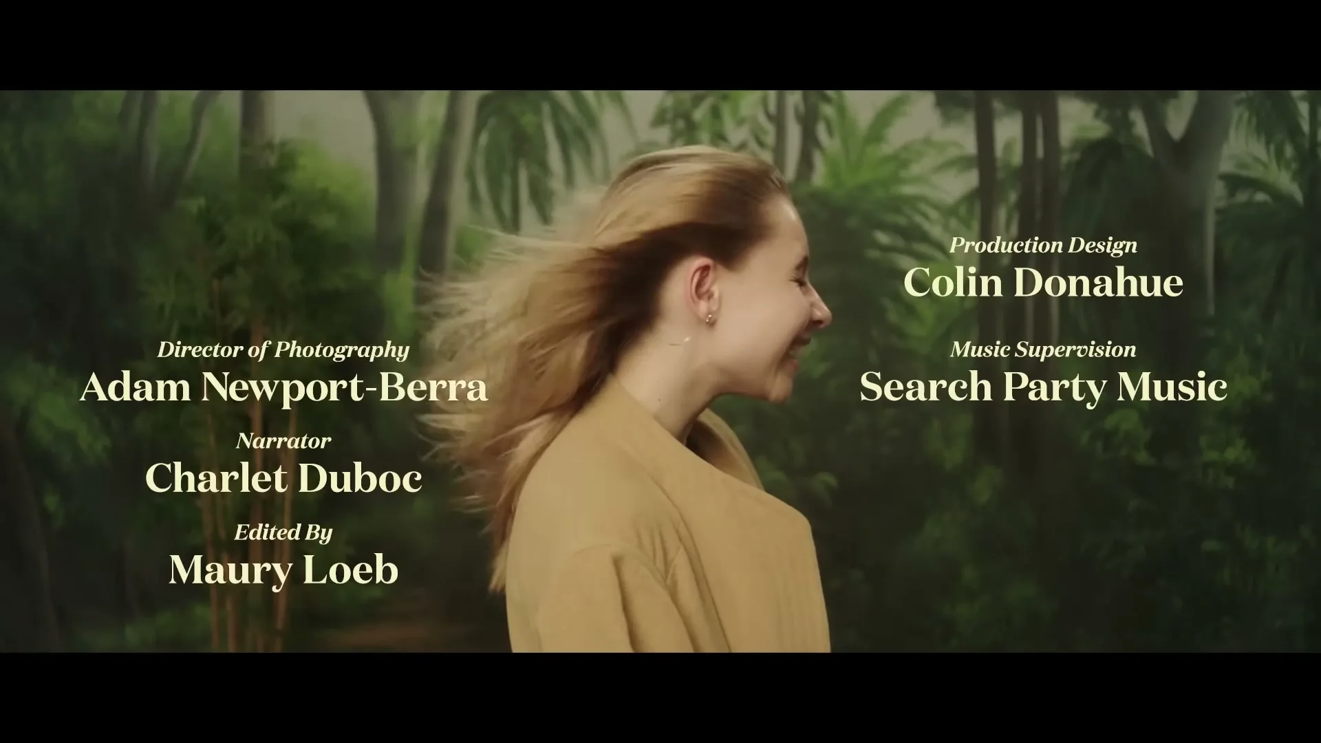 A young woman with blonde hair, wearing a tan coat, smiles as her hair blows in the wind, with production credits overlaid on a jungle-like background.