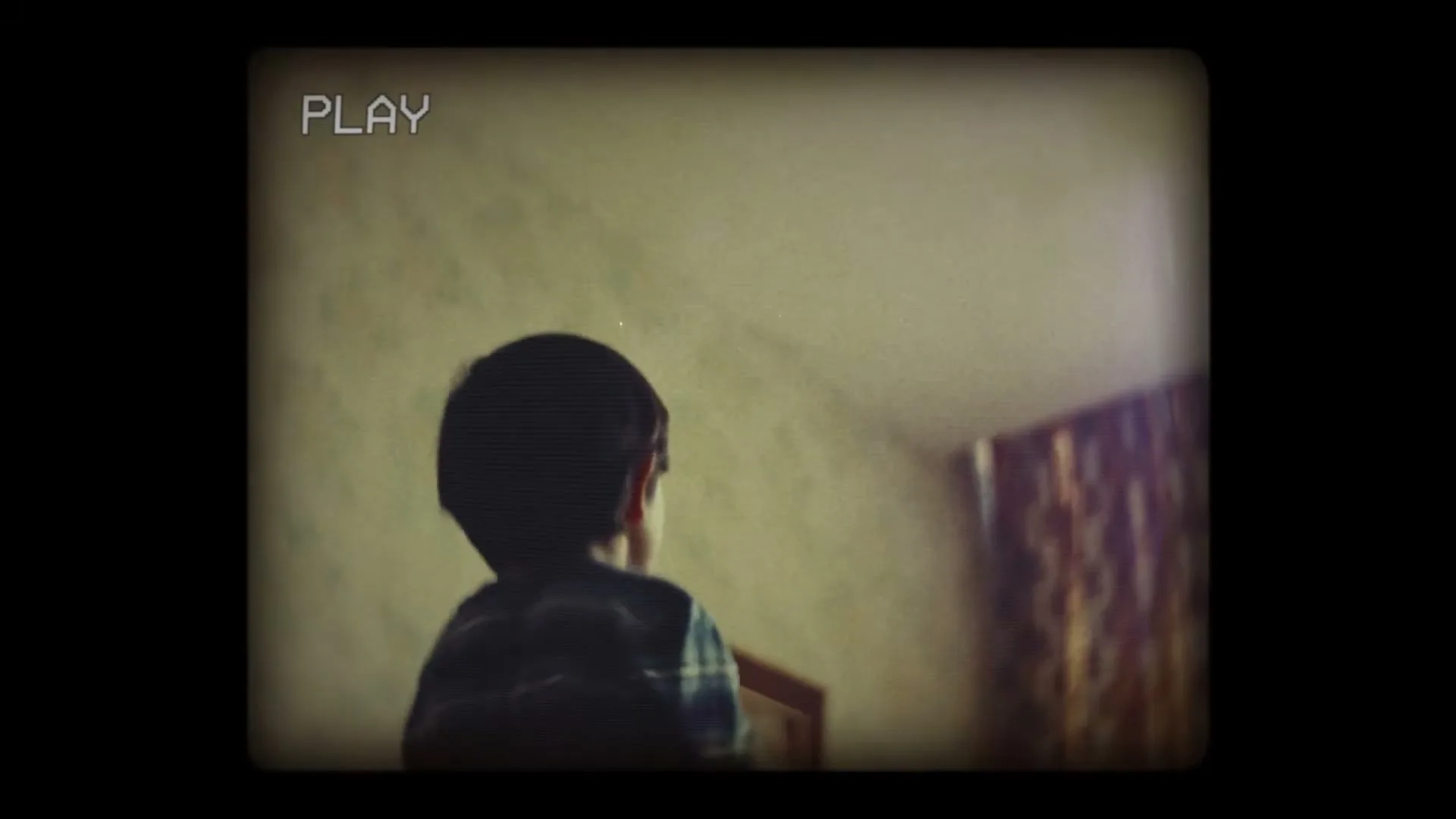 A young boy, seen from behind, plays with a toy airplane, moving it through the air in a dimly lit room, captured with a vintage camcorder effect.
