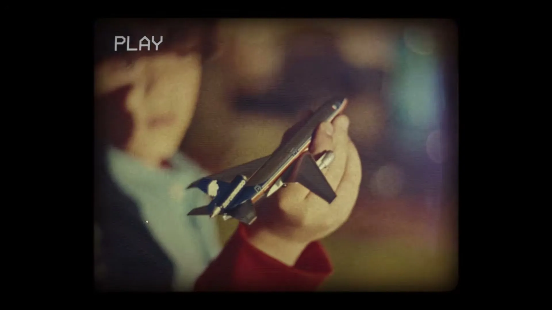 A child's hand holds a toy airplane, with the child's face blurred in the background, evoking a nostalgic, home video feel.