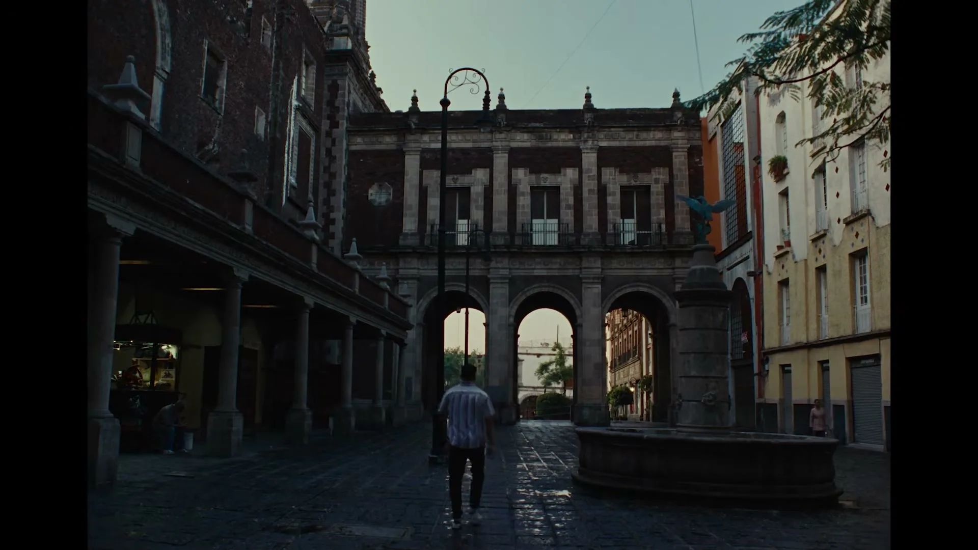A man in a striped shirt walks away from the camera down a cobblestone street towards a large building with archways in a historic town square.