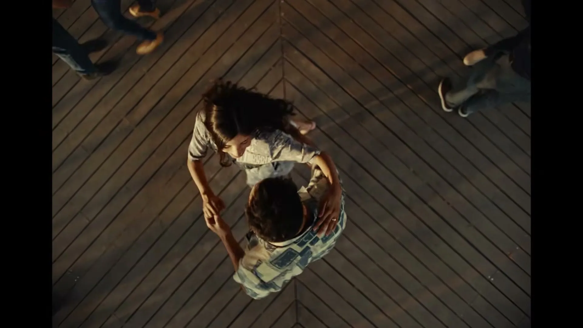 A young couple dances closely on a wooden floor, seen from directly above, with other people's legs visible around them.