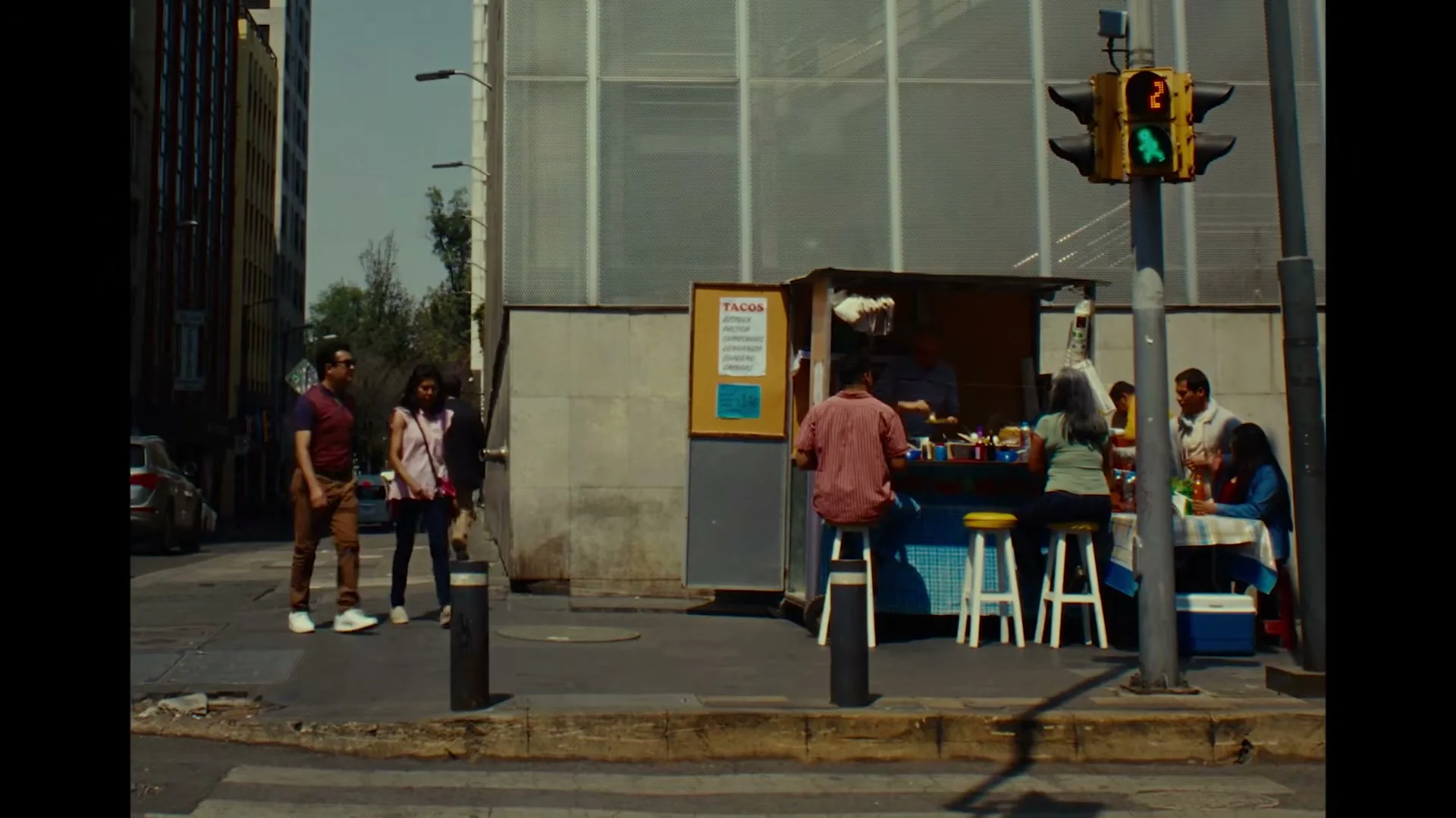People walk past a street food stall with customers eating tacos on a sunny day in a bustling city.