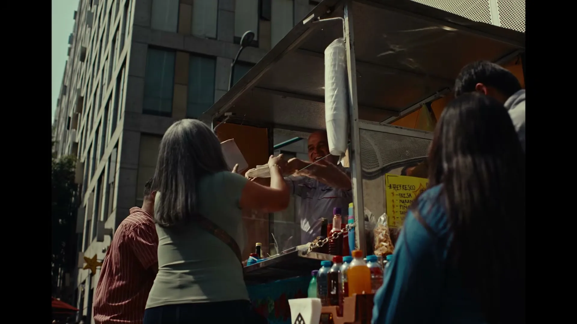 A vendor in a food cart serves a woman with gray hair, while other customers wait in a bustling city street.