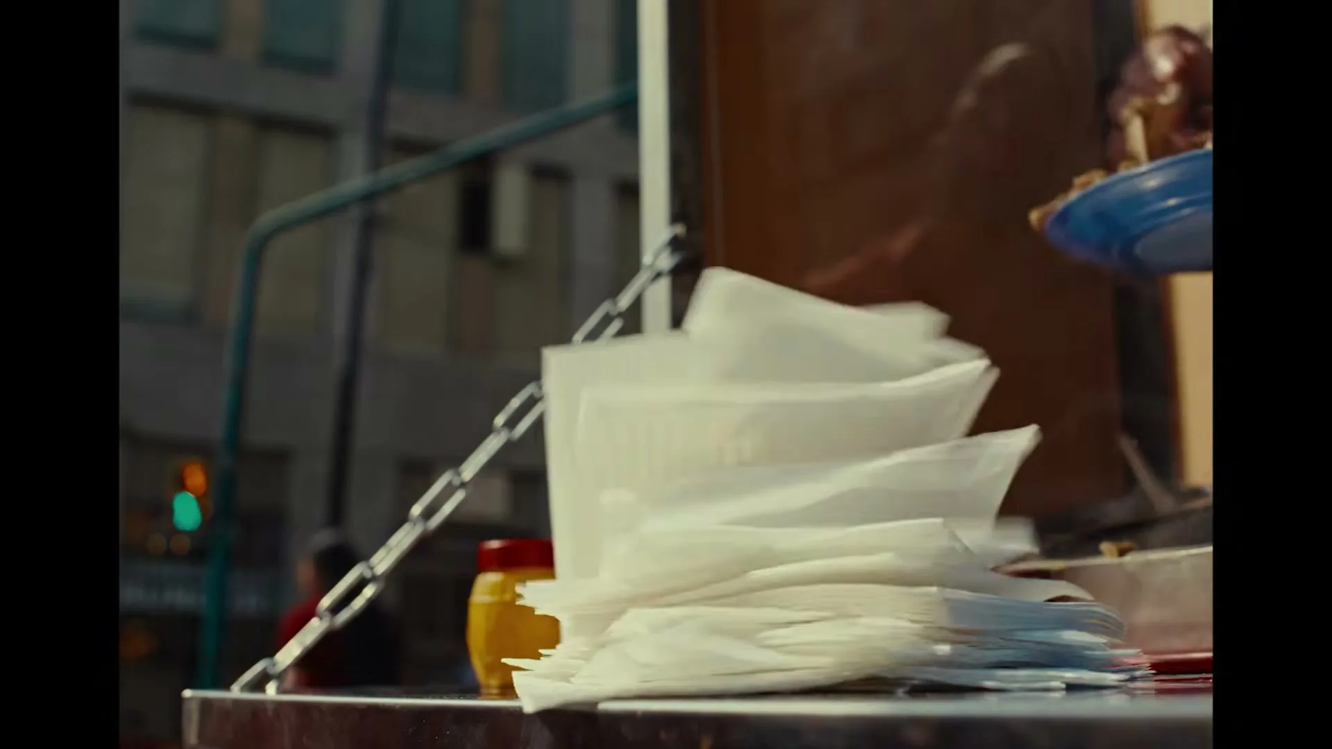 A stack of white napkins on a street food cart is blown by wind, with a yellow mustard bottle and city buildings in the background.