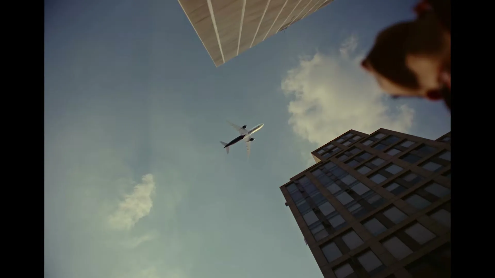 A low-angle shot captures a white airplane flying across a blue sky, framed by two tall buildings and a person's head in the foreground.