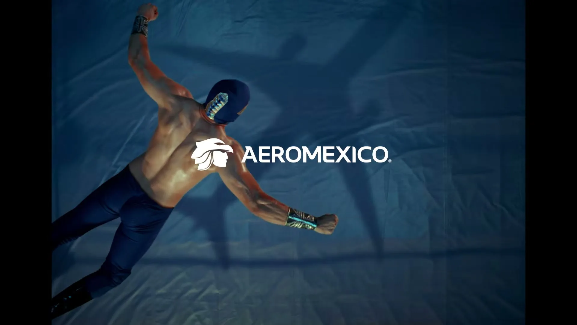 A male luchador in a blue mask and trunks, with a bare chest, is seen from above, lying on a blue wrestling mat, with the Aeromexico logo and text appearing on screen.
