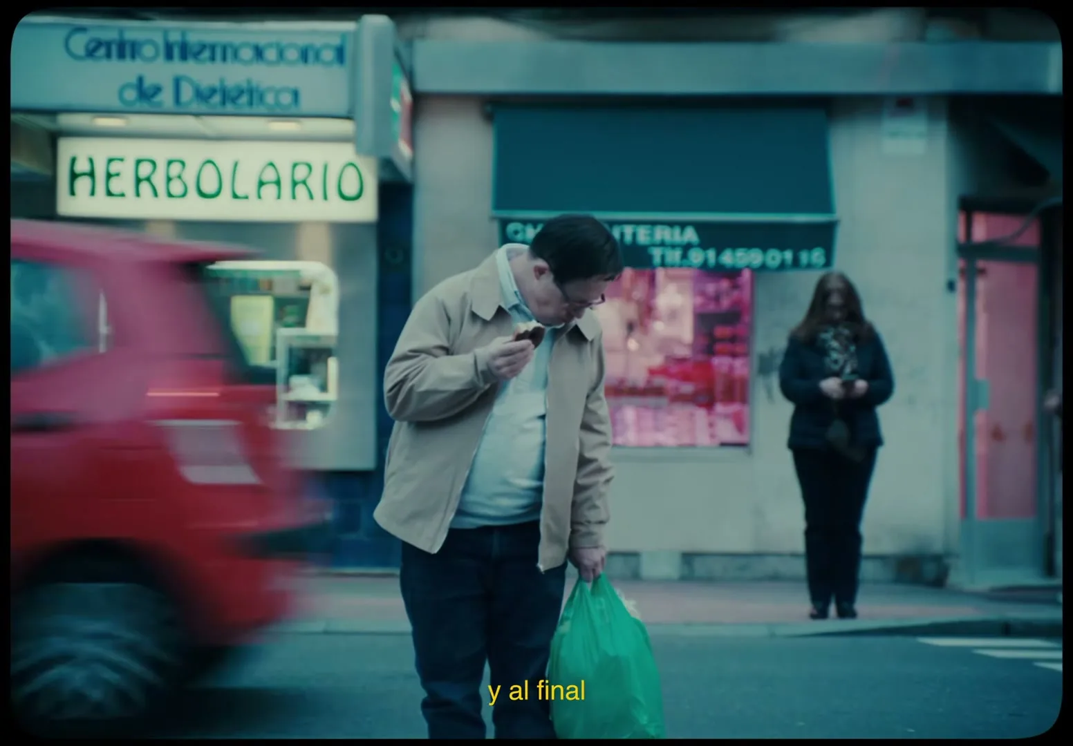 A man with Down syndrome stands in the middle of a crosswalk, eating a chocolate donut and holding a green shopping bag, as people and cars pass by on a city street.