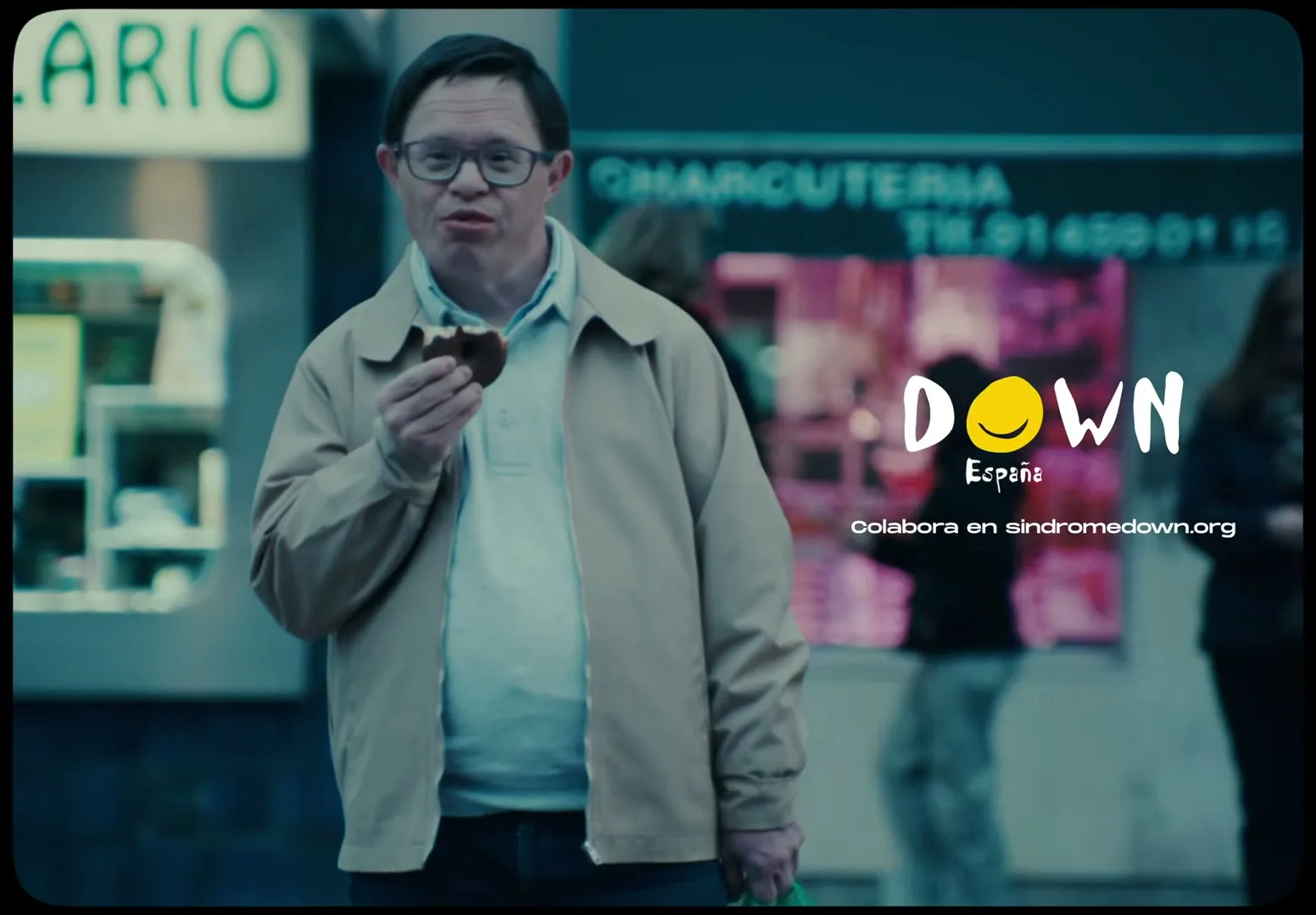 A man with Down syndrome, wearing glasses and a beige jacket, stands on a city street, holding a half-eaten cookie and a green bag.