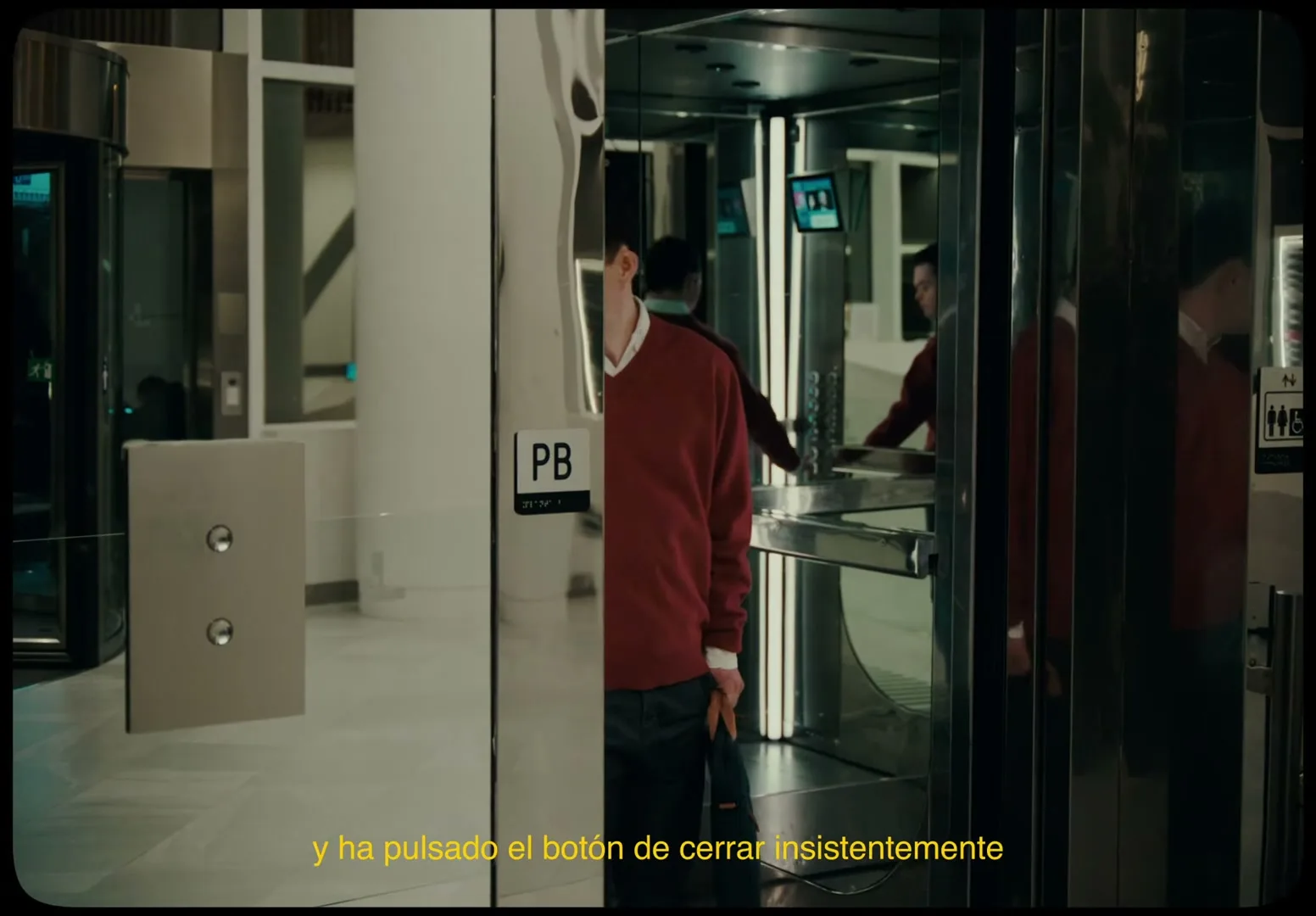 A young man in a red sweater and white collared shirt stands in front of an elevator, looking up and to the right, in a modern building lobby.
