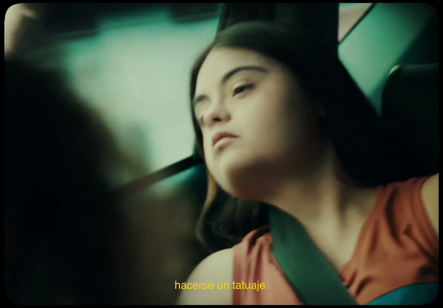 A young female with Down syndrome, wearing a seatbelt and an orange shirt, looks out the window of a moving car, with a blurred person in the foreground.